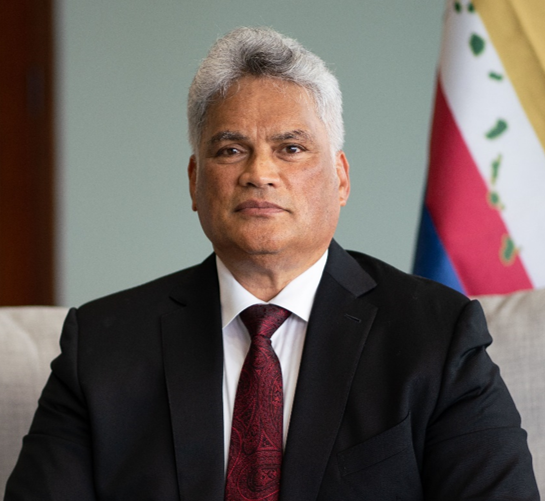 King Edmund Keliʻi Paki-Silva Jr. seated in a suit and tie with the Hawaiian flag in the background, representing his role as Sovereign of the Kingdom of Hawaiʻi.