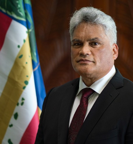 King Edmund Keliʻi Paki-Silva Jr. in a suit and tie, standing before the Hawaiian flag, representing the Kingdom of Hawaiʻi.