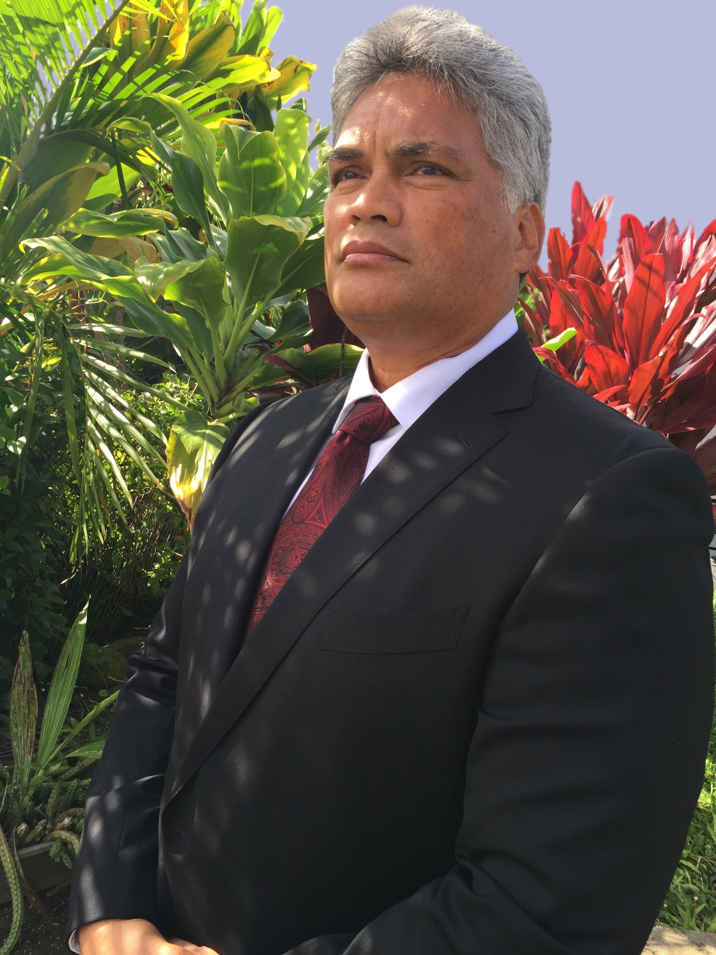 King Edmund Keliʻi Paki-Silva Jr. wearing a black suit and red tie, standing outdoors in front of tropical plants, looking forward with a dignified expression.