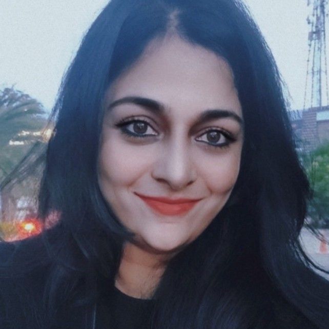 A smiling woman with long black hair, wearing a dark top, photographed outdoors with a soft background.