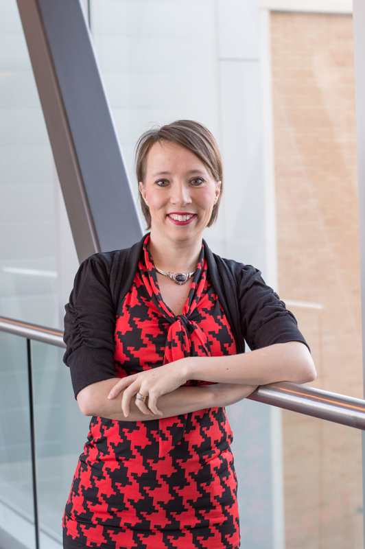 A woman wearing a red and black patterned dress with a black cardigan, smiling while leaning against a railing indoors.