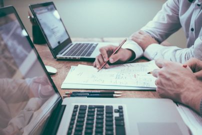 Two people working together at a desk with laptops and documents, discussing financial plans and making notes with pencils.