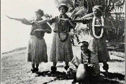 Black and white photograph of Hawaiian dancers in traditional attire performing hula, with a kupuna seated in front holding a gourd drum.