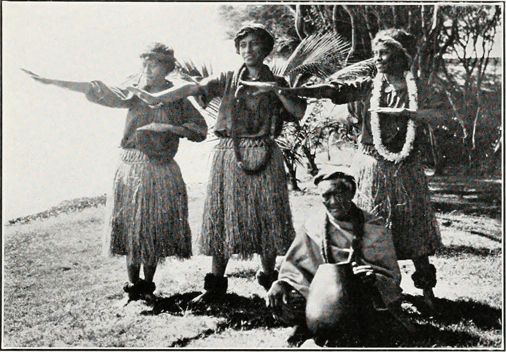 Black and white photograph of Hawaiian dancers in traditional attire performing hula, with a kupuna seated in front holding a gourd drum.