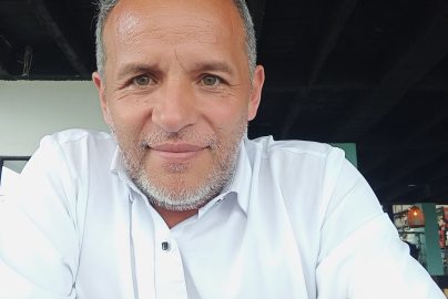 A man in a white shirt smiling warmly at the camera, seated indoors with natural light.