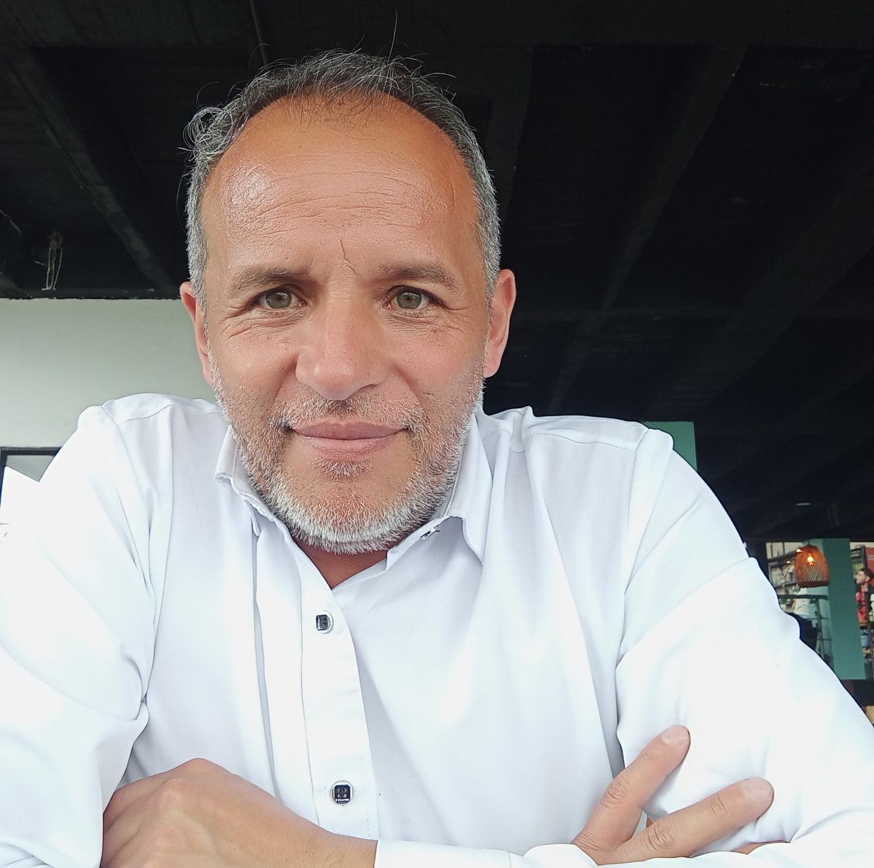 A man in a white shirt smiling warmly at the camera, seated indoors with natural light.
