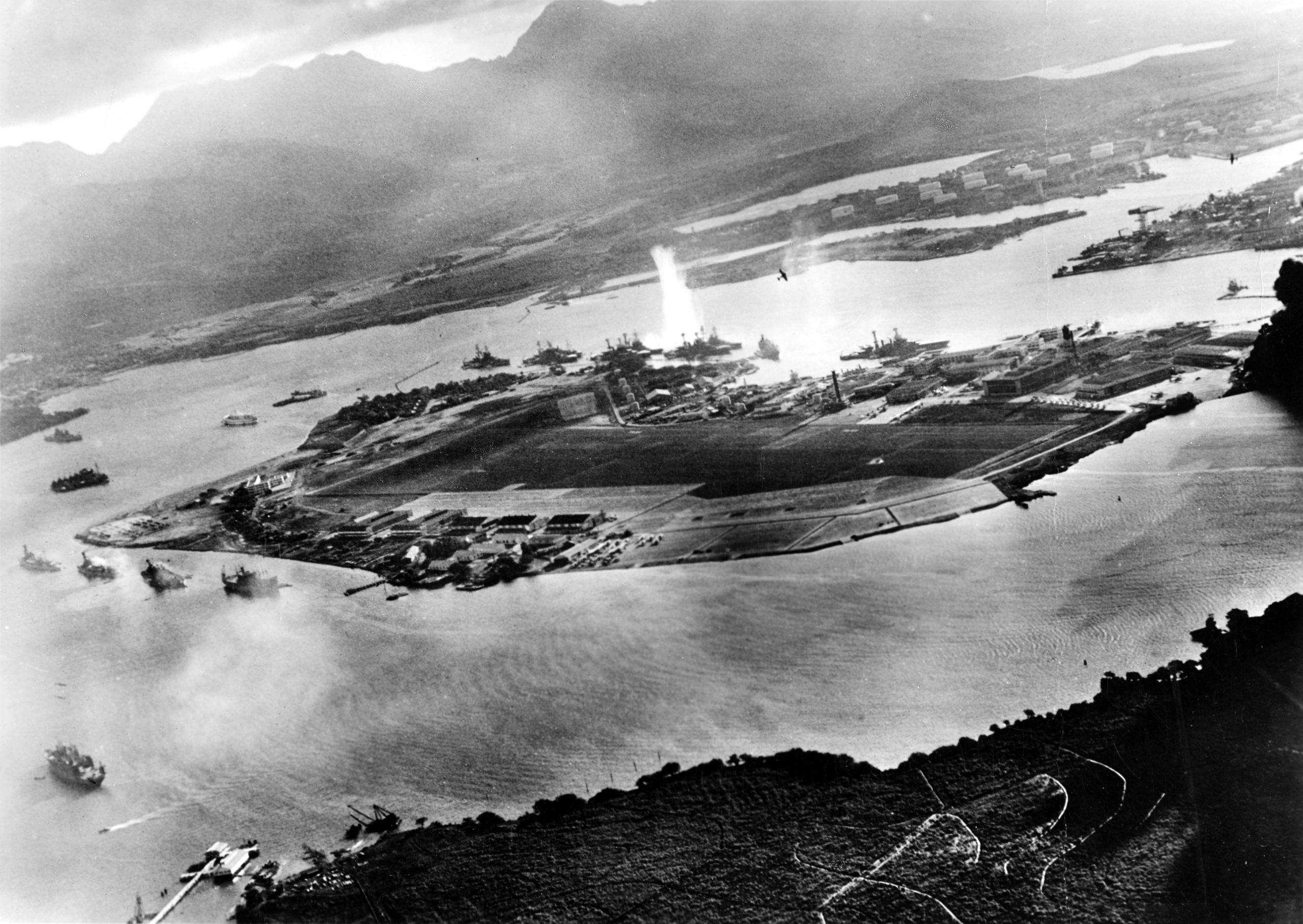 Photograph taken from a Japanese plane during the torpedo attack on ships moored on both sides of Ford Island. View looks about east, with the supply depot, submarine base and fuel tank farm in the right center distance. A torpedo has just hit USS West Virginia on the far side of Ford Island (center). Other battleships moored nearby are (from left): Nevada, Arizona, Tennessee (inboard of West Virginia), Oklahoma (torpedoed and listing) alongside Maryland, and California. On the near side of Ford Island, to the left, are light cruisers Detroit and Raleigh, target and training ship Utah and seaplane tender Tangier. Raleigh and Utah have been torpedoed, and Utah is listing sharply to port. Japanese planes are visible in the right center (over Ford Island) and over the Navy Yard at right. Japanese writing in the lower right states that the photograph was reproduced by authorization of the Navy Ministry. U.S. Naval History and Heritage Command Photograph. {{brizy_dc_image_alt imageSrc=