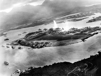 Photograph taken from a Japanese plane during the torpedo attack on ships moored on both sides of Ford Island. View looks about east, with the supply depot, submarine base and fuel tank farm in the right center distance. A torpedo has just hit USS West Virginia on the far side of Ford Island (center). Other battleships moored nearby are (from left): Nevada, Arizona, Tennessee (inboard of West Virginia), Oklahoma (torpedoed and listing) alongside Maryland, and California. On the near side of Ford Island, to the left, are light cruisers Detroit and Raleigh, target and training ship Utah and seaplane tender Tangier. Raleigh and Utah have been torpedoed, and Utah is listing sharply to port. Japanese planes are visible in the right center (over Ford Island) and over the Navy Yard at right. Japanese writing in the lower right states that the photograph was reproduced by authorization of the Navy Ministry. U.S. Naval History and Heritage Command Photograph.