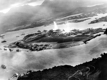 Photograph taken from a Japanese plane during the torpedo attack on ships moored on both sides of Ford Island. View looks about east, with the supply depot, submarine base and fuel tank farm in the right center distance. A torpedo has just hit USS West Virginia on the far side of Ford Island (center). Other battleships moored nearby are (from left): Nevada, Arizona, Tennessee (inboard of West Virginia), Oklahoma (torpedoed and listing) alongside Maryland, and California. On the near side of Ford Island, to the left, are light cruisers Detroit and Raleigh, target and training ship Utah and seaplane tender Tangier. Raleigh and Utah have been torpedoed, and Utah is listing sharply to port. Japanese planes are visible in the right center (over Ford Island) and over the Navy Yard at right. Japanese writing in the lower right states that the photograph was reproduced by authorization of the Navy Ministry. U.S. Naval History and Heritage Command Photograph. {{brizy_dc_image_alt entityId=