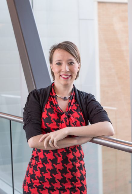 A woman wearing a red and black patterned dress with a black cardigan, smiling while leaning against a railing indoors. {{brizy_dc_image_alt imageSrc=