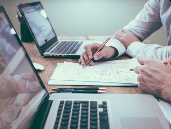 Two people working together at a desk with laptops and documents, discussing financial plans and making notes with pencils.