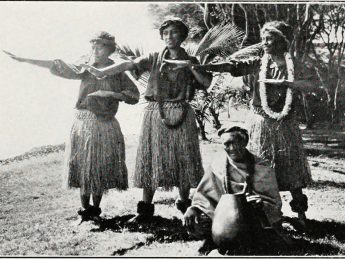 Black and white photograph of Hawaiian dancers in traditional attire performing hula, with a kupuna seated in front holding a gourd drum.