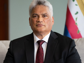 King Edmund Keliʻi Paki-Silva Jr. seated in a suit and tie with the Hawaiian flag in the background, representing his role as Sovereign of the Kingdom of Hawaiʻi.