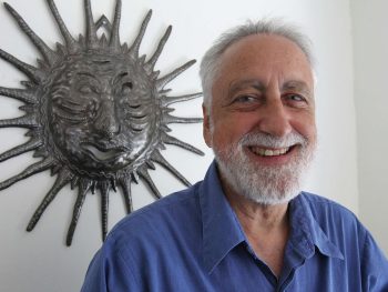 Portrait of Lanny Sinkin, Aliʻi Manaʻo Nui (Chief Advisor) of the Kingdom of Hawaiʻi, smiling in front of a sun sculpture, symbolizing wisdom and guidance.